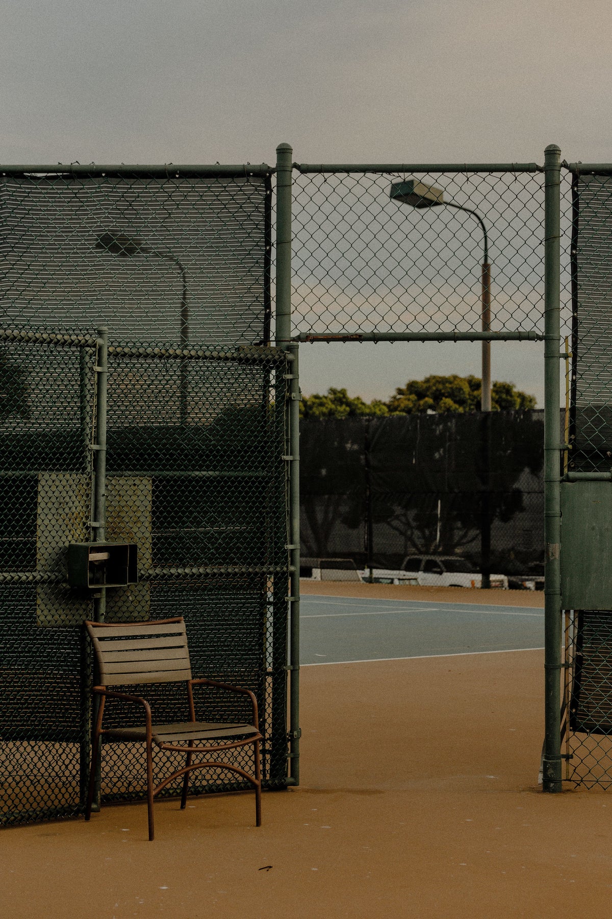 Outdoor tennis court viewed through an open gate, with floodlight poles and an umpire chair